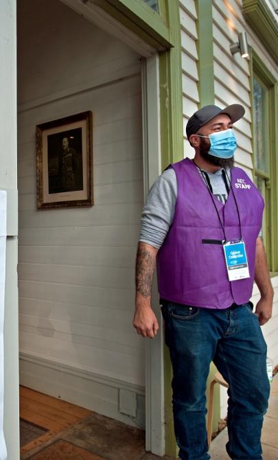 Man wearing face mask and purple vest standing new a doorway. On the wall around the door are instructions on how to vote in a federal election.